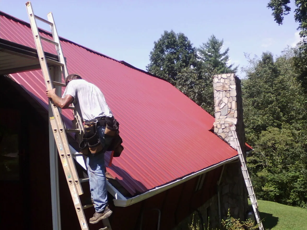 Roofer on a ladder beside a red metal roof and stone chimney — Soffit & Fascia Repair by Roofing Brothers in Mebane